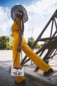 Fashionable woman posing outdoors in bright yellow jumpsuit with striking striped hat and sunglasses.