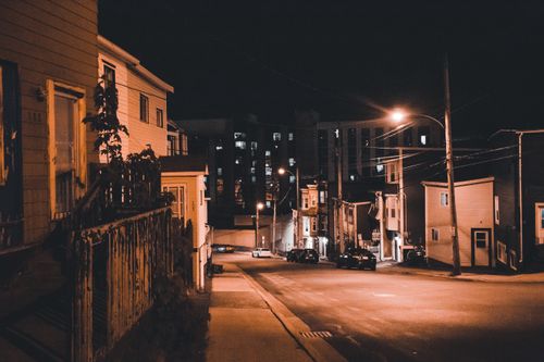 Free A deserted urban street at night illuminated by warm streetlights with parked cars and buildings. Stock Photo