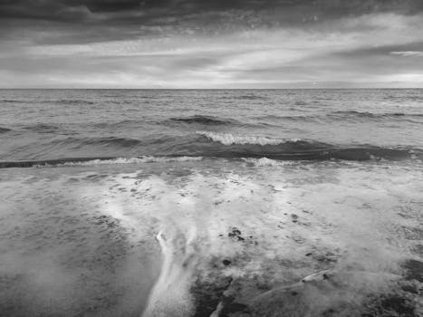 Grayscale image of ocean waves gently crashing onto the tranquil shore under a cloudy sky.