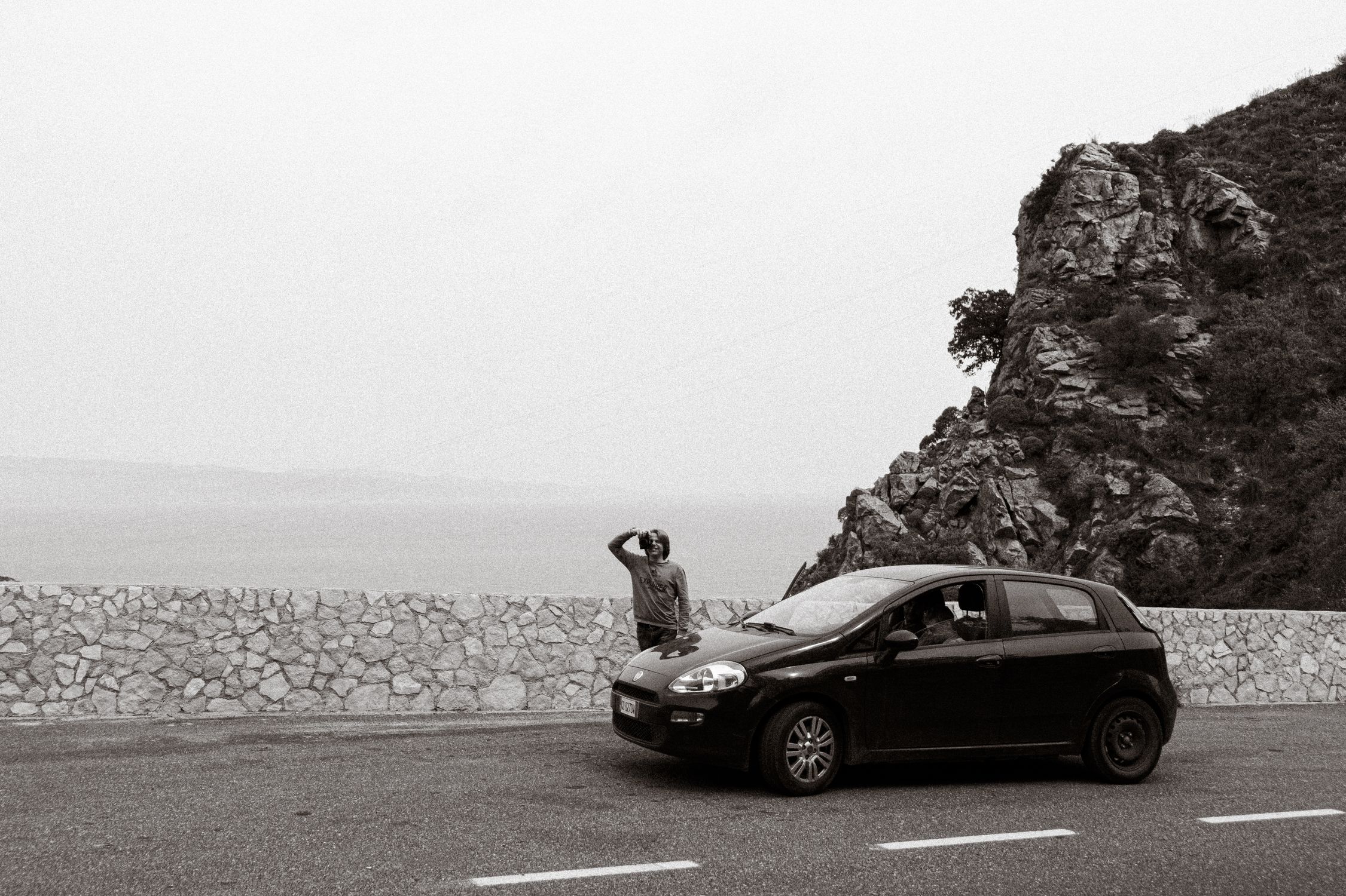 Black car with a person standing by a rocky mountain roadside in grayscale.