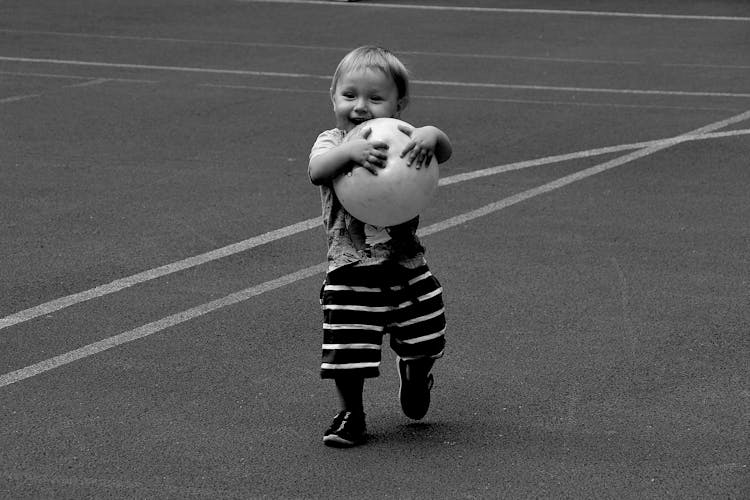 Grayscale Photo Of A Happy Toddler Holding A Ball