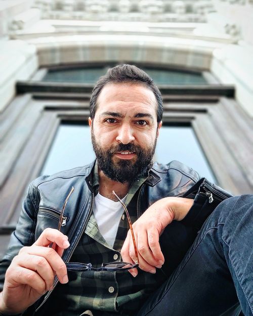 Free Bearded man in a leather jacket holding glasses, sitting outdoors. Stock Photo