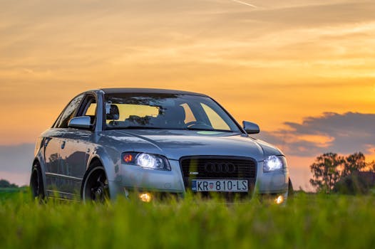 Close-up of a parked Audi car with headlights on during sunset in a grassy outdoor setting.