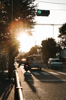 Cars and motorcycles drive on a city road, silhouetted by the rising sun.