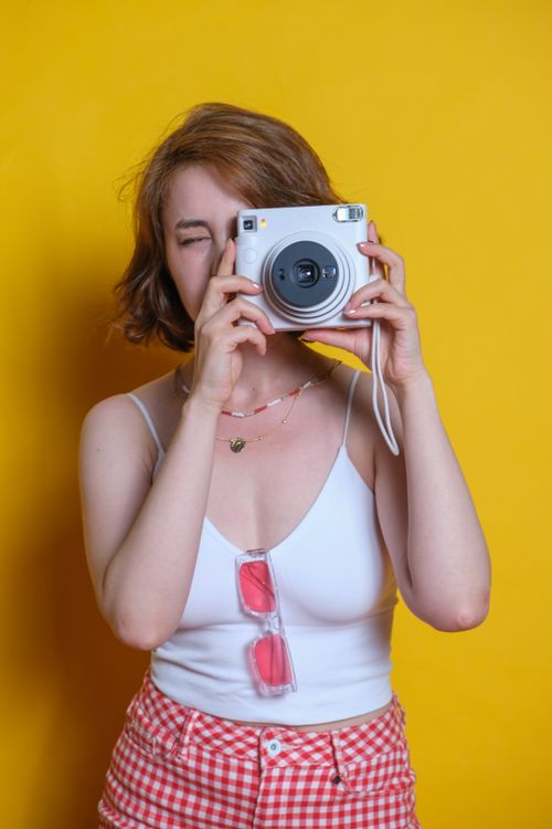 Free A young woman in a spaghetti strap top takes a photo with an instant camera against a yellow background. Stock Photo