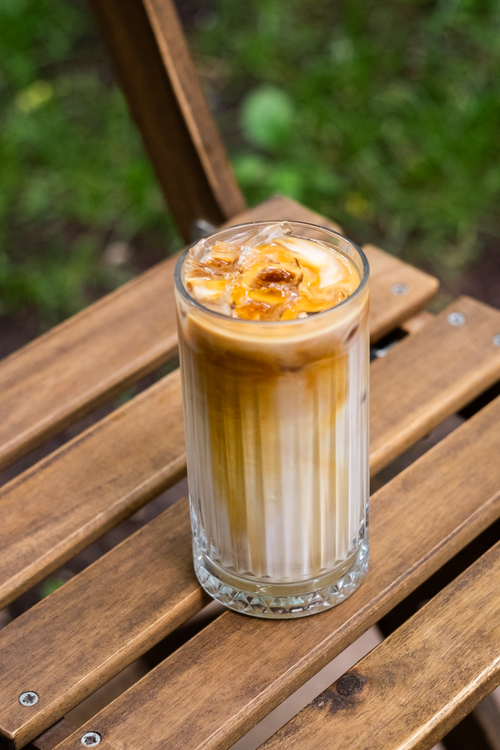 Free A tall glass of iced coffee sits on a wooden chair, set against a garden backdrop, evoking a cool and serene atmosphere. Stock Photo