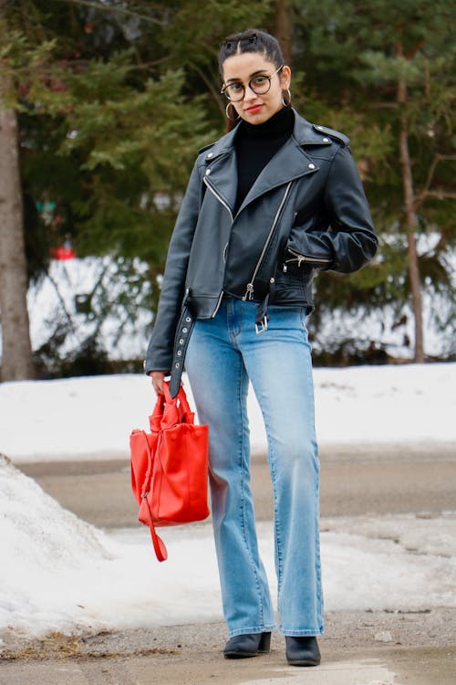 A Woman in Black Leather and Denim Jeans Holding Her Bag while Standing on the Street