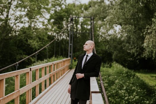 Bald man in a black coat stands thoughtfully on a wooden bridge amidst greenery.