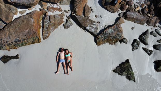 An aerial shot of a couple lying on a beach surrounded by rocks, exuding relaxation and vacation vibes.