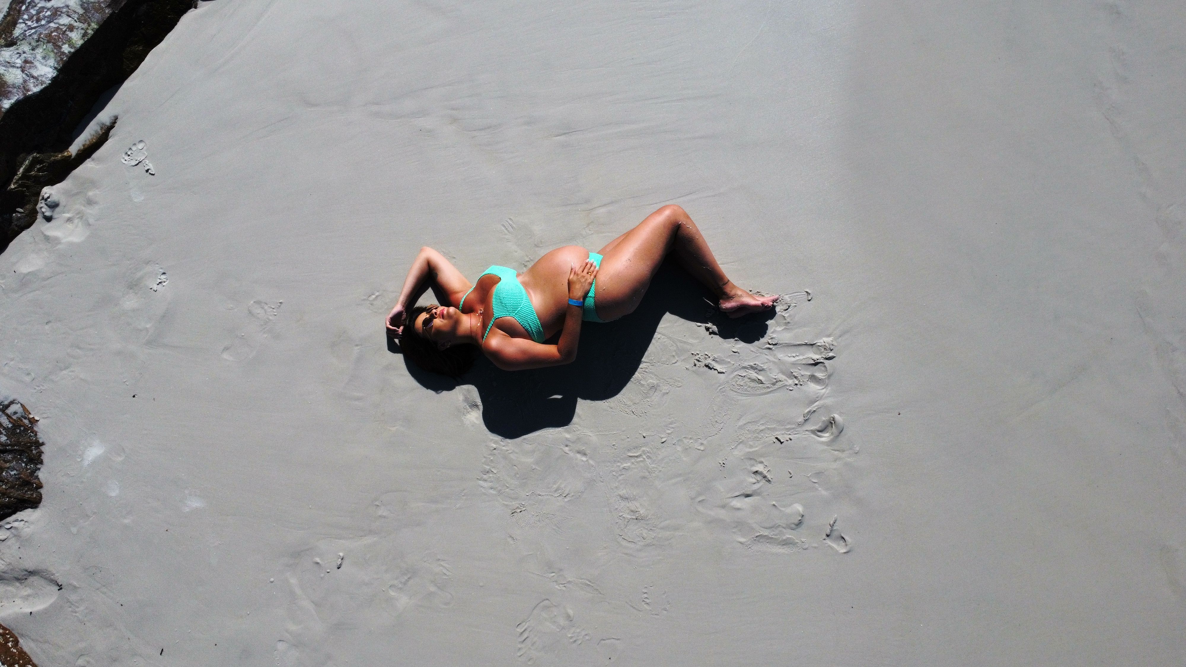 Aerial view of a pregnant woman sunbathing on a sandy beach wearing sunglasses and swimwear.