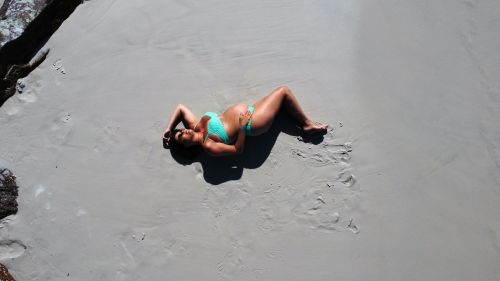 Free Aerial view of a pregnant woman sunbathing on a sandy beach wearing sunglasses and swimwear. Stock Photo