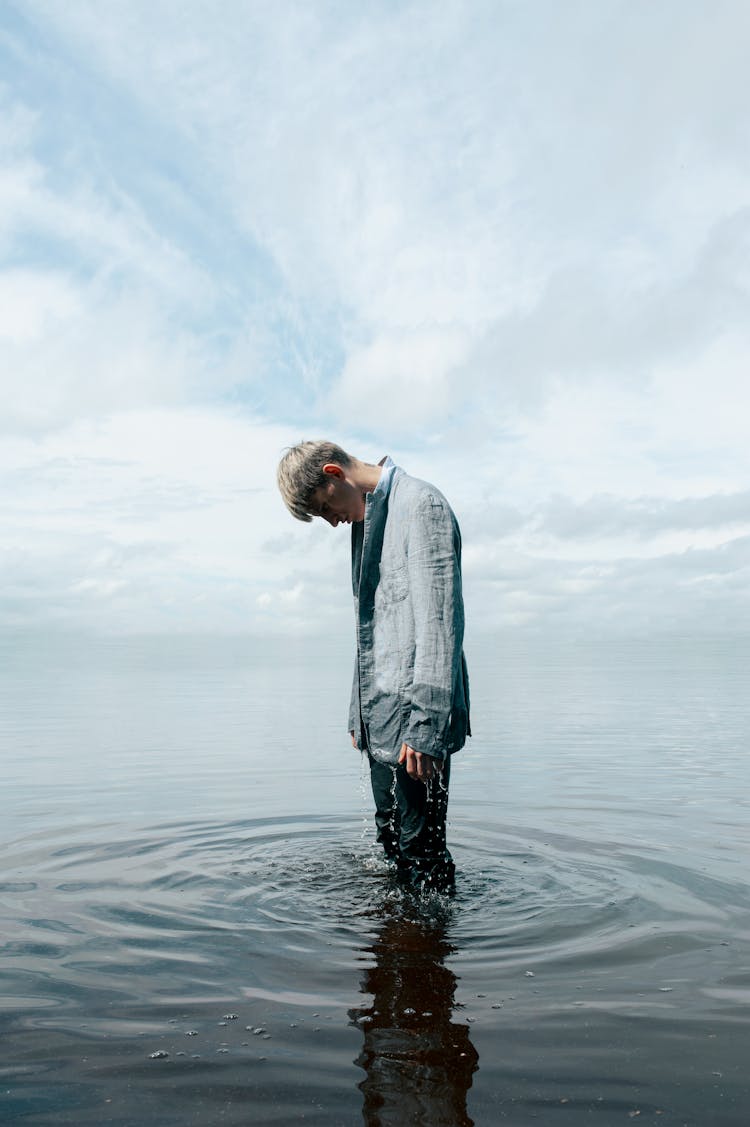 Fully Clothed Man Standing Knee Deep In Water With His Head Down 