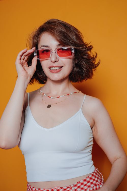 Free Confident woman wearing stylish red sunglasses and white top, set against a vibrant orange backdrop. Stock Photo