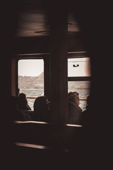 Silhouetted ferry passengers gaze through windows with a serene sea view.