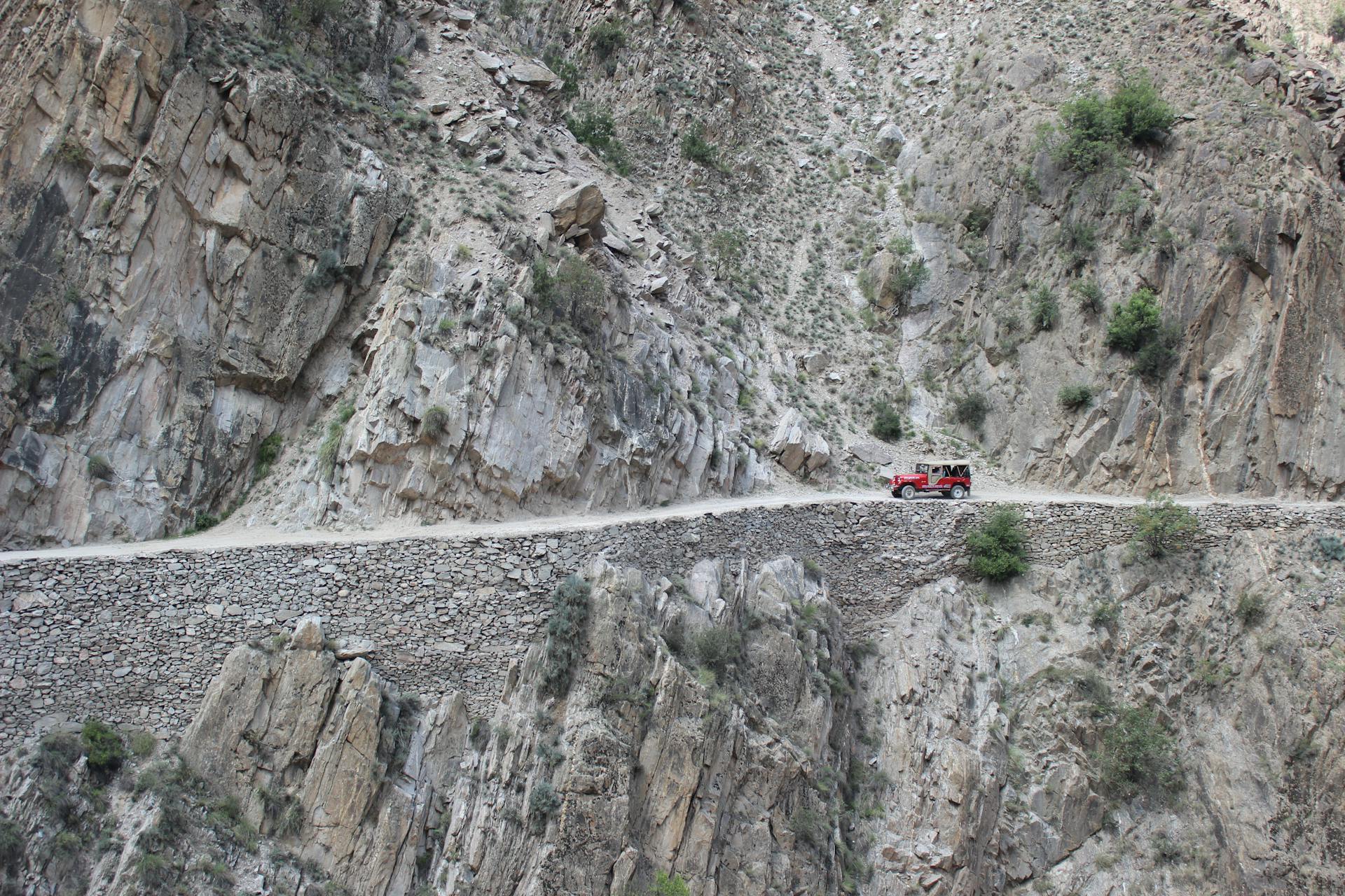 Red Jeep navigating a narrow cliffside road in dramatic mountain landscape near Skardu, Pakistan.