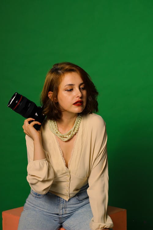 Woman in Beige Long Sleeve Shirt Holding Black Camera in Green Background 