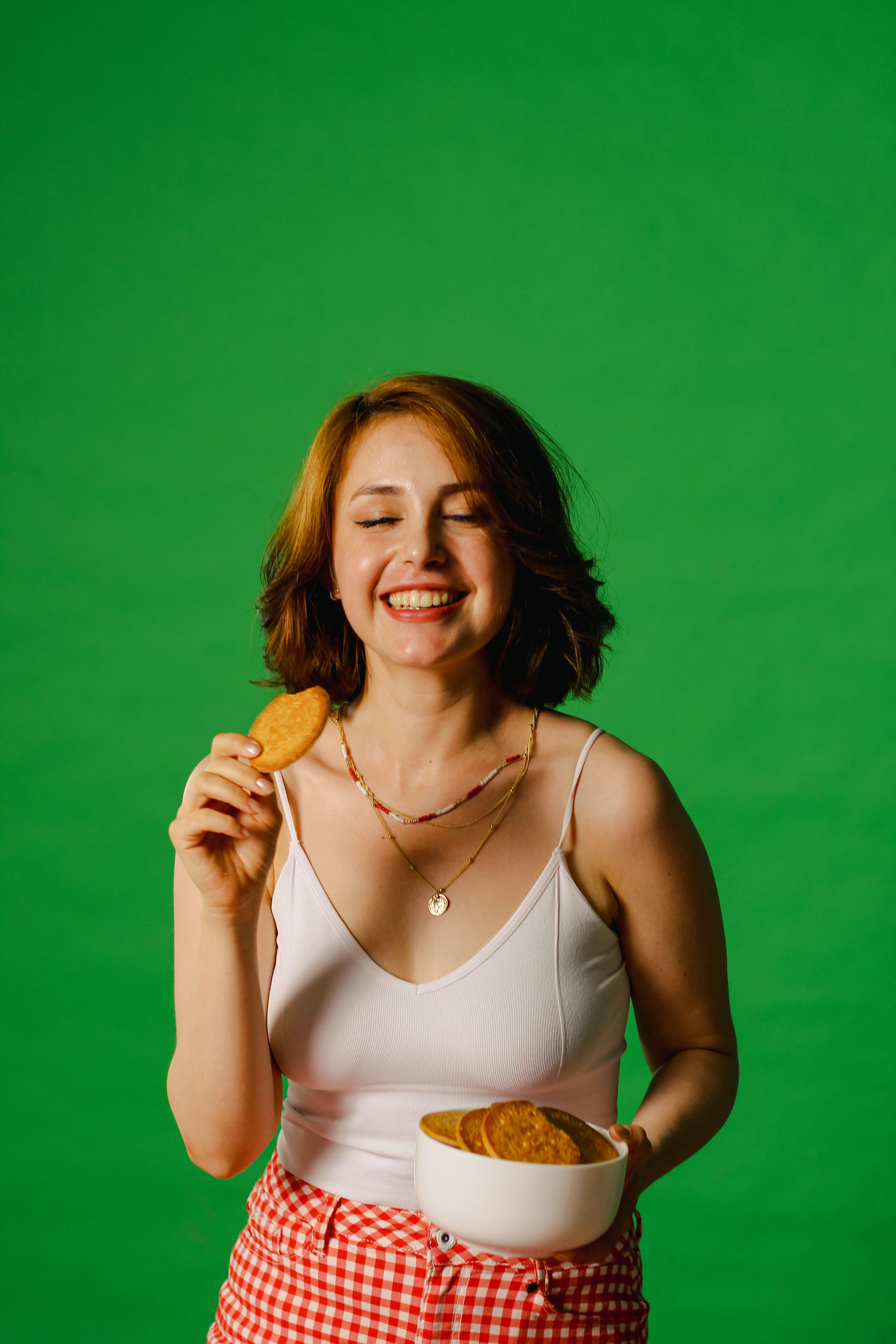 Cheerful woman with closed eyes holding a cookie and bowl in front of a vibrant green backdrop.
