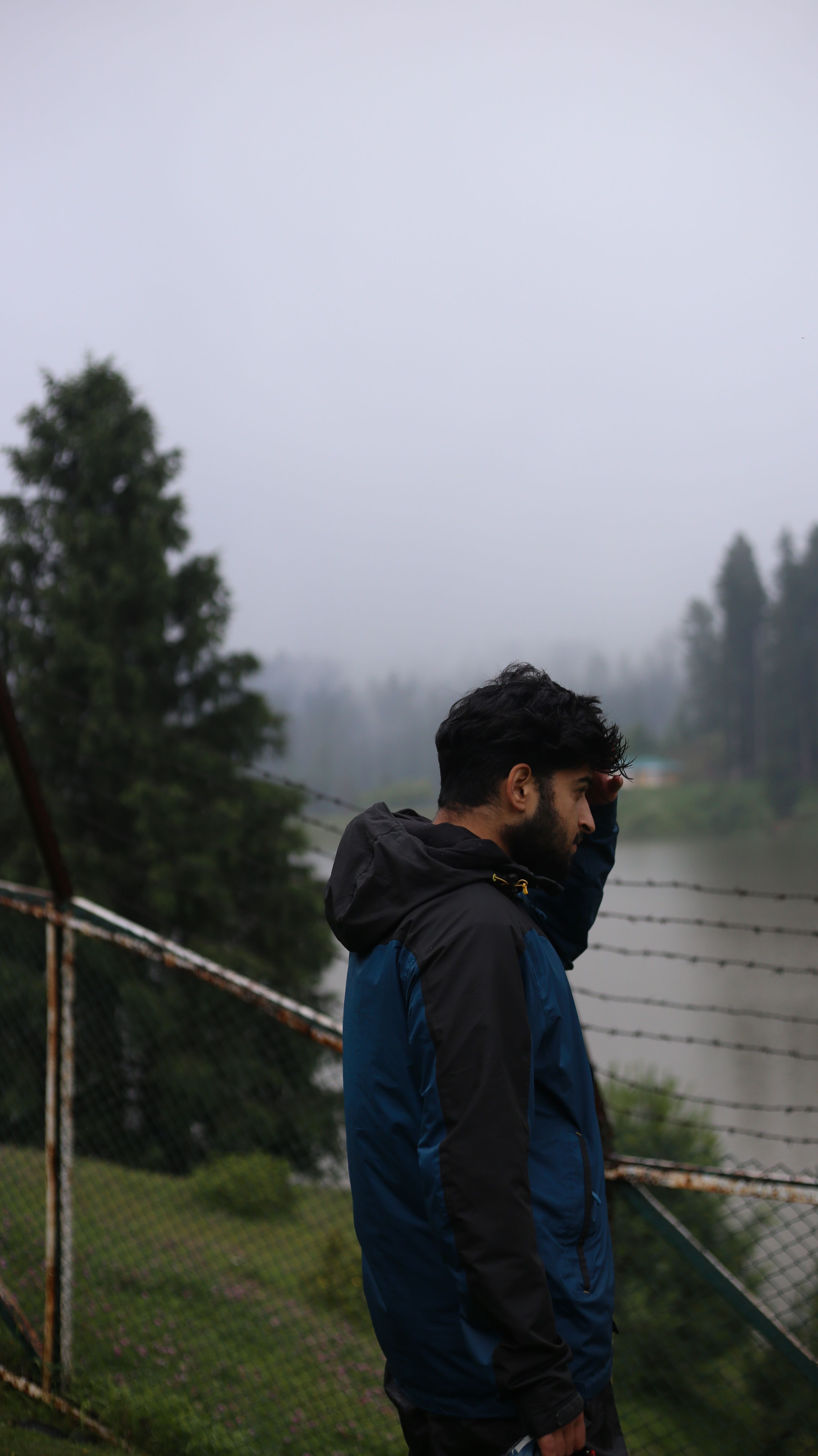 A man in a blue jacket stands by a misty lake on a cloudy day, contemplating the serene view.