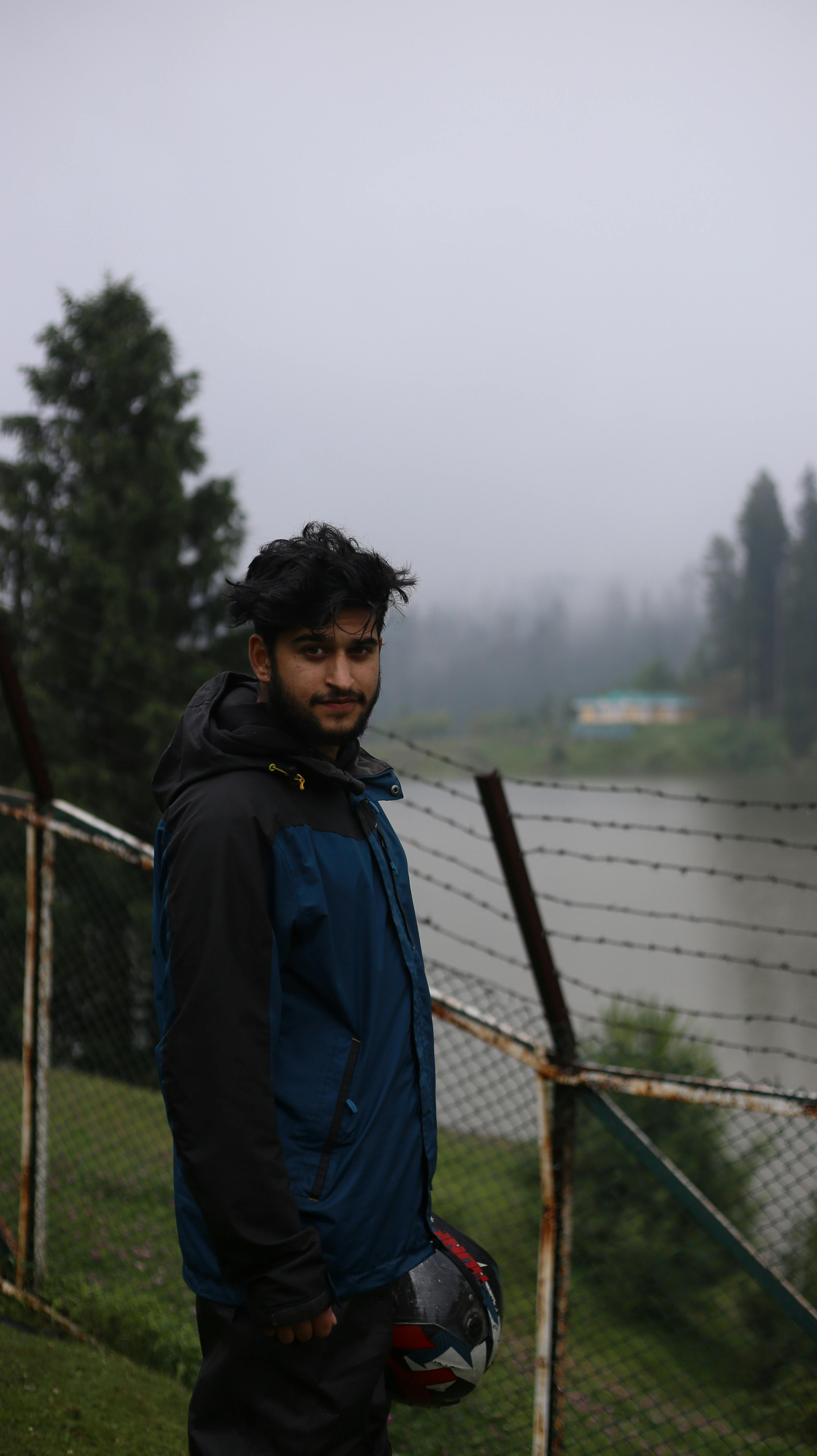 Young man wearing jacket holding helmet by a lakeside fence with misty trees in the background.