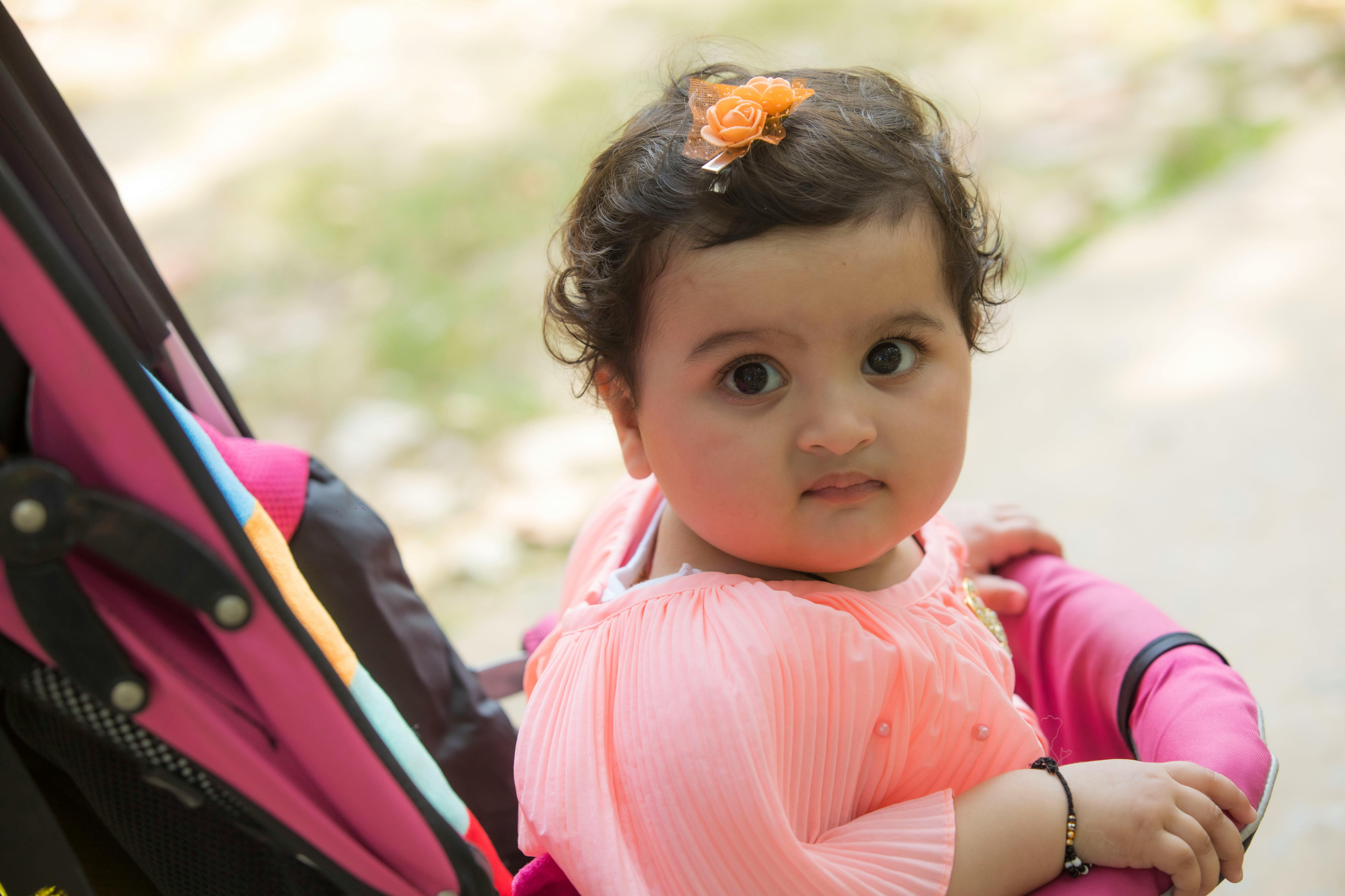 Charming baby girl with curly hair sitting in a stroller outdoors on a sunny day, looking curious.
