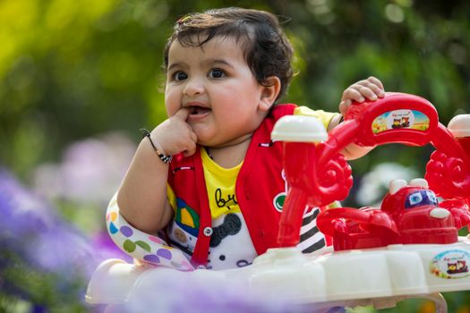 Cute baby seated in a colorful stroller, playfully putting finger in the mouth outdoors.
