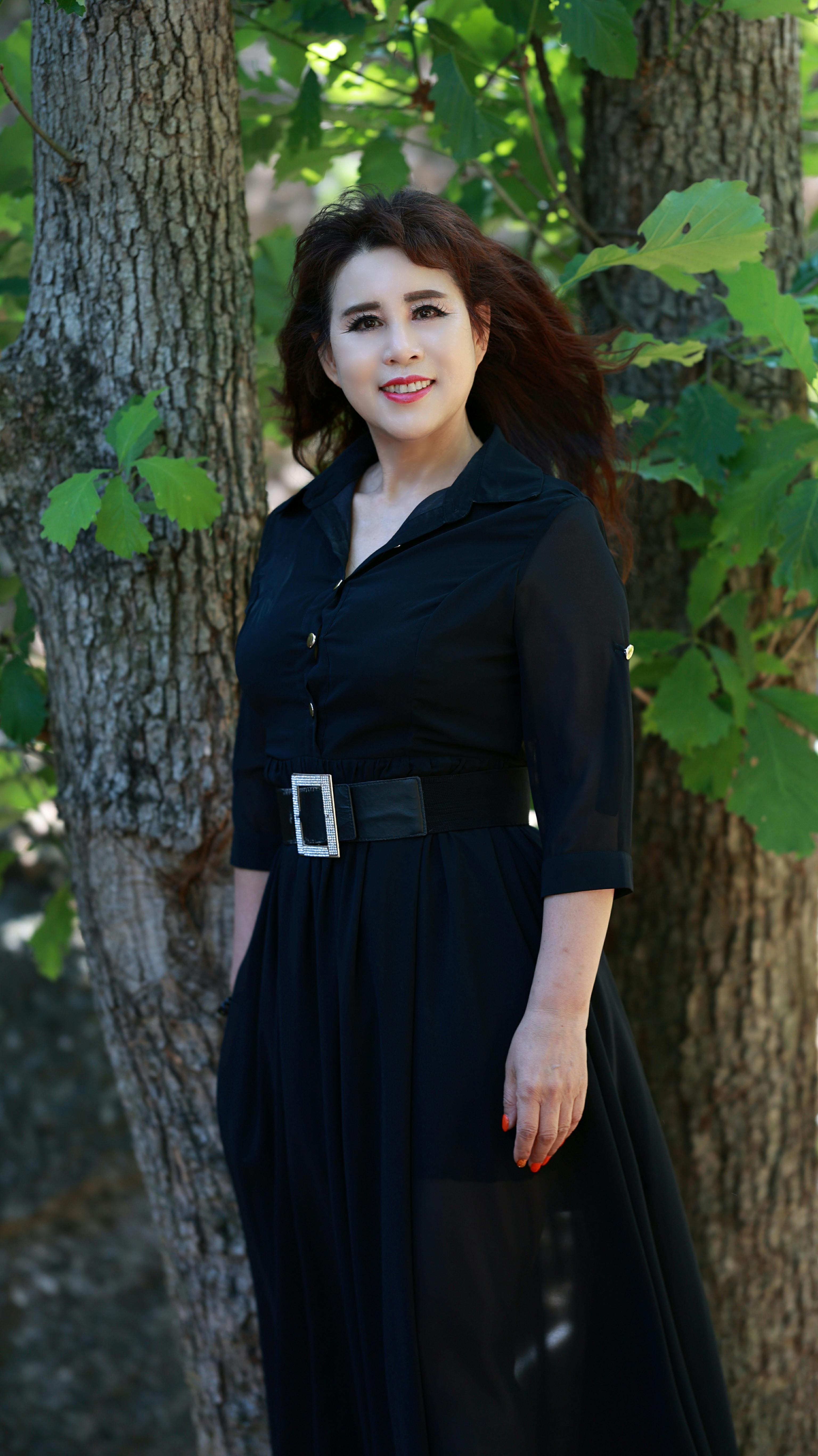 Stylish woman standing by trees, wearing a chic black dress. Natural beauty captured in outdoor setting.
