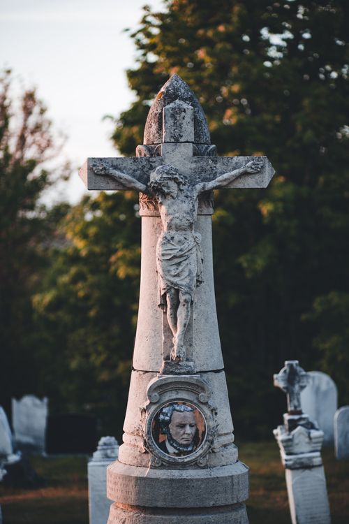 Free A cross-adorned gravestone in a historic cemetery surrounded by trees. Stock Photo