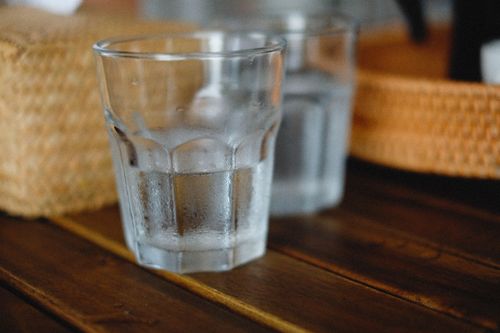 Free A cold, refreshing glass of water on a rustic wooden table, with woven baskets in the background. Stock Photo