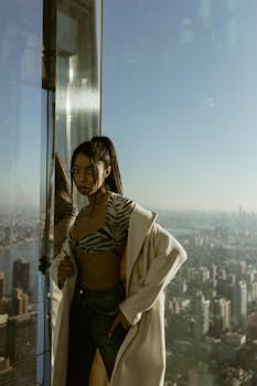 Fashionable woman poses by the window with a stunning view of New York City skyline.