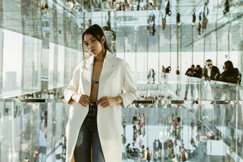 Free Young woman in a white coat poses confidently inside a modern glass structure reflecting New York City's skyline. Stock Photo