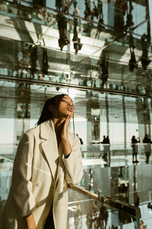 Free Fashionable woman in beige coat poses amid reflective glass panels, New York. Stock Photo