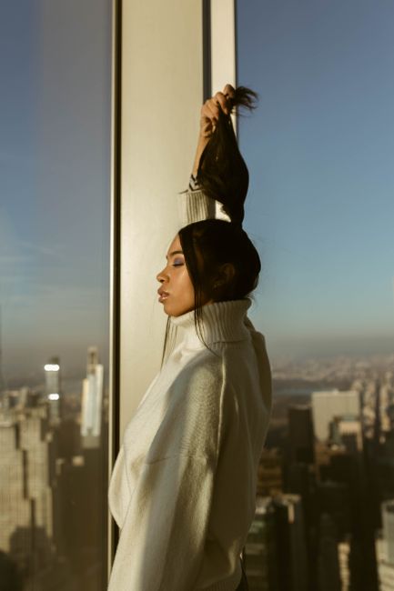 Elegant woman in white sweater posing by the window of a New York City skyscraper.