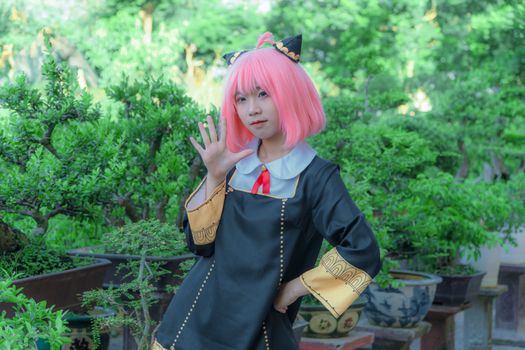 Cosplayer in a pink wig and costume poses outdoors surrounded by bonsai plants.