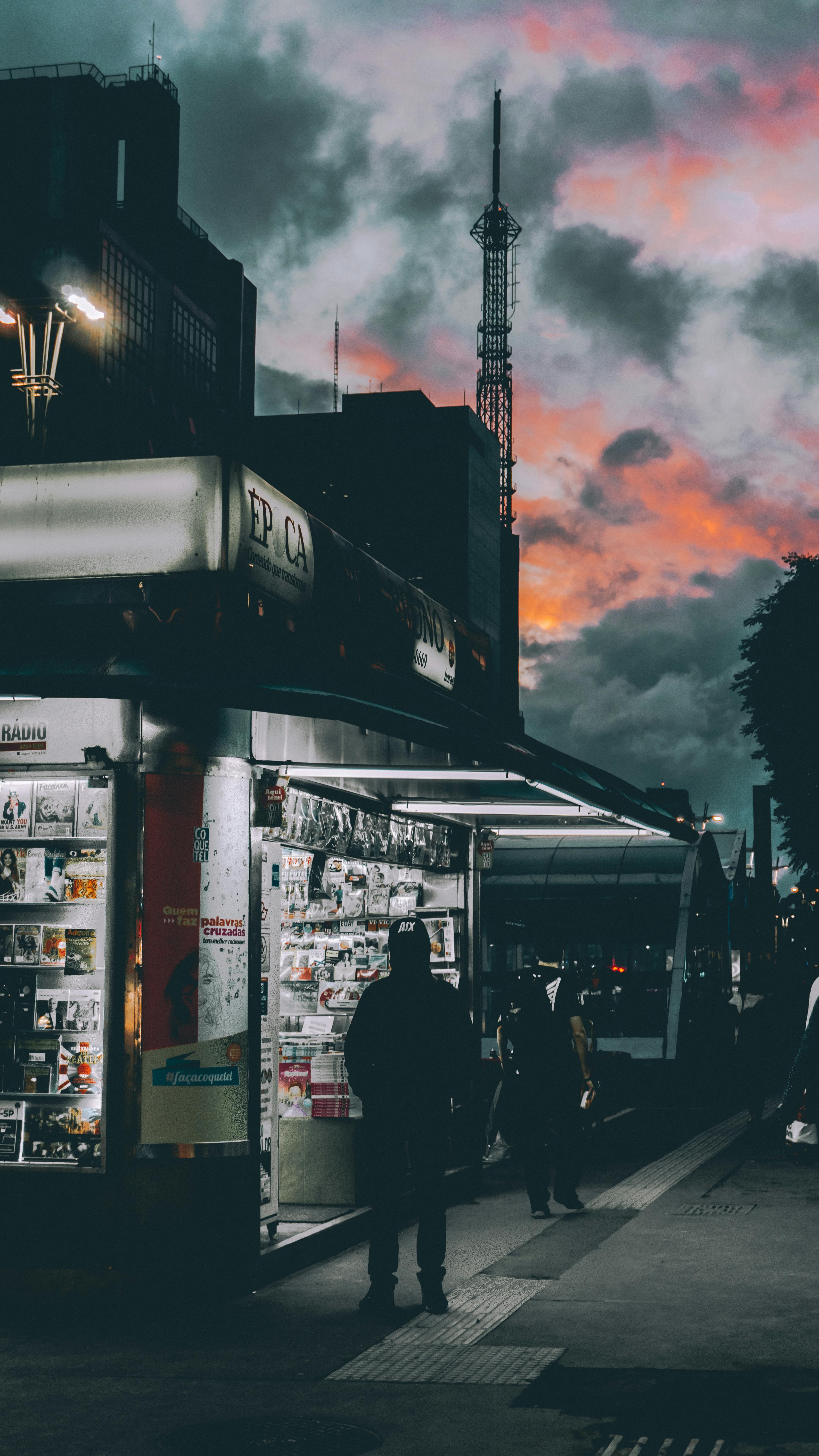 在戏剧性的天空下，繁忙的城市街道旁，热闹的报摊与迷人的黄昏构成了一幅生动的场景