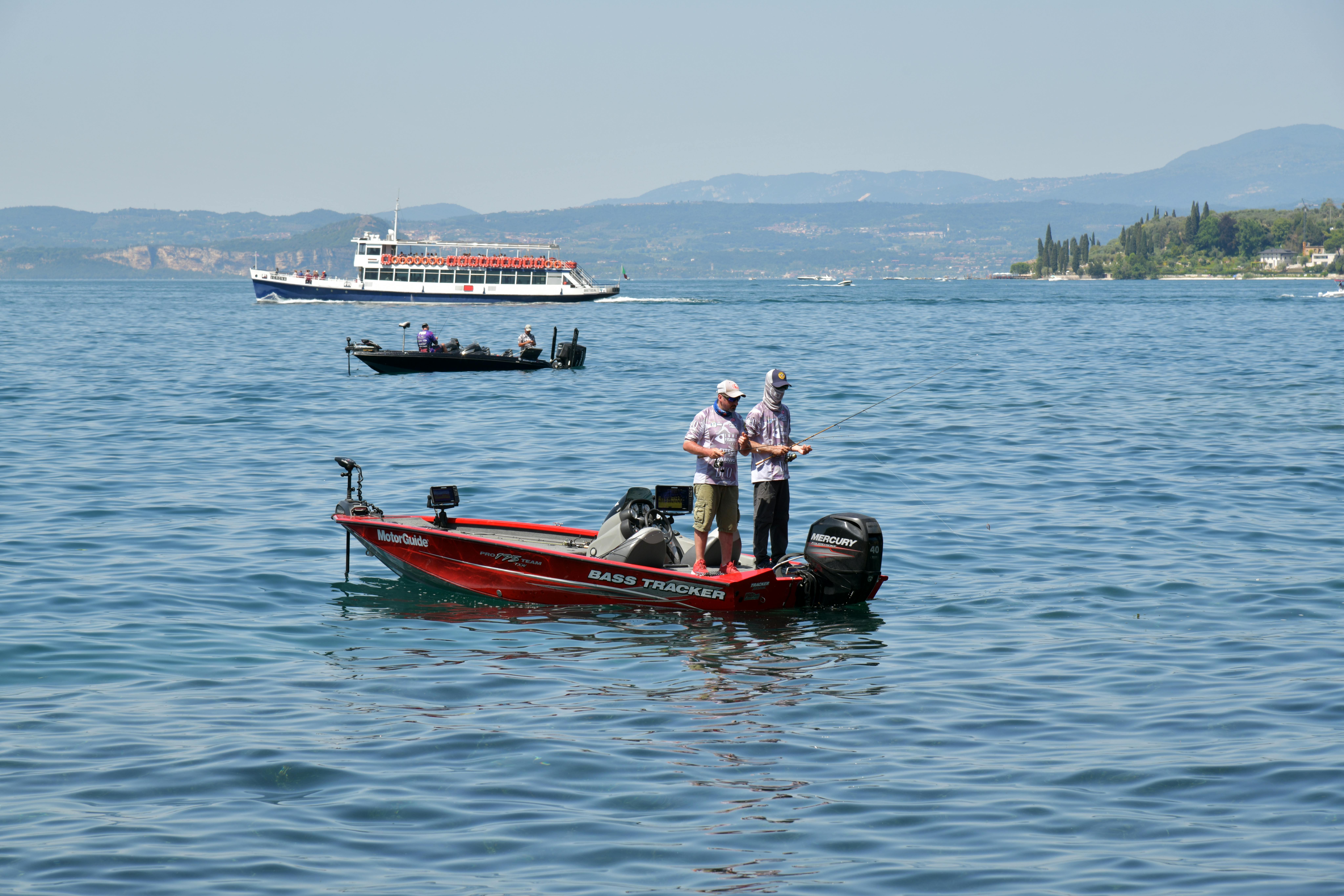 Two men fishing on Lake Garda, Italy, with boats and scenic mountain views.