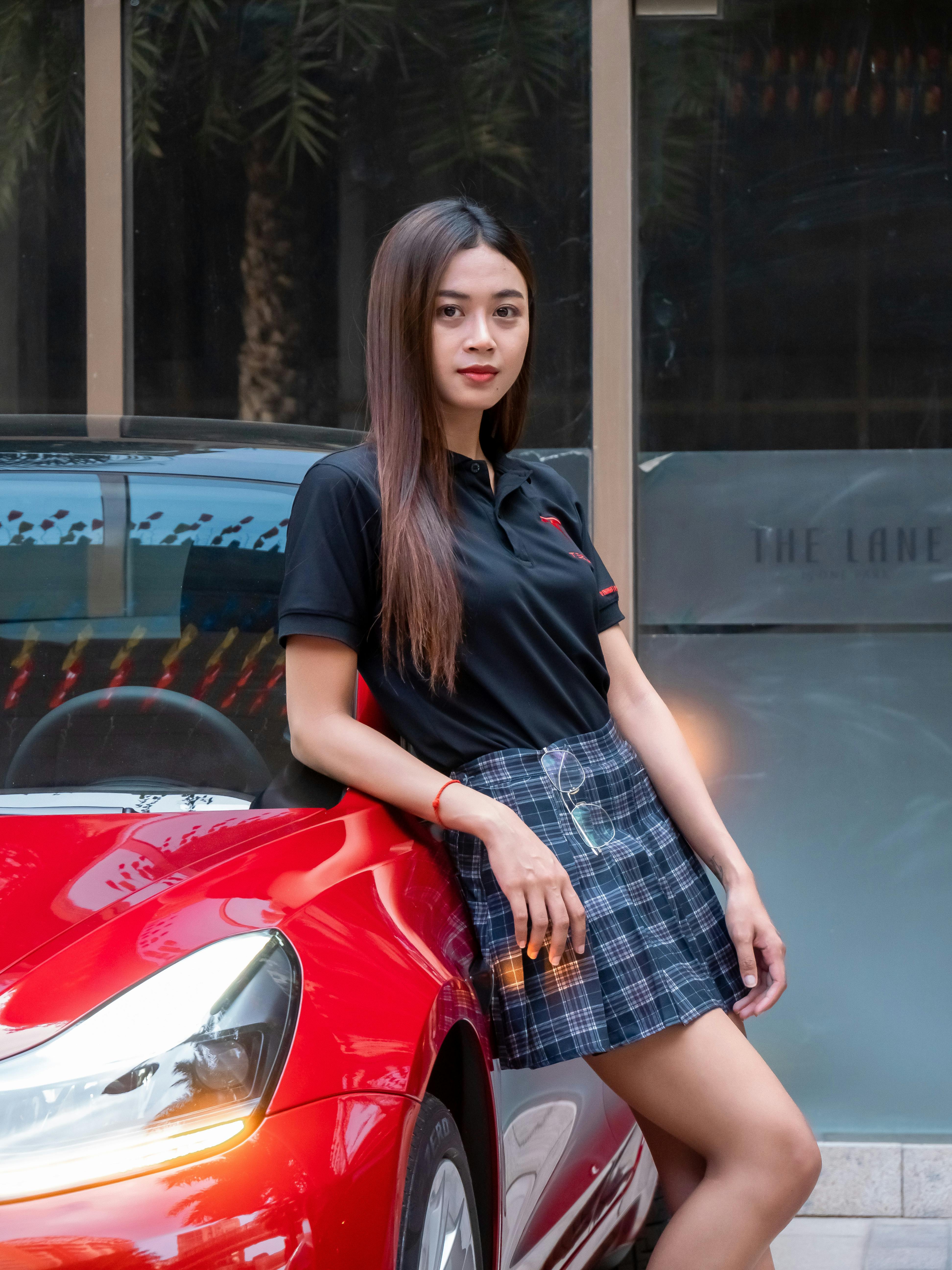 Free A stylish Asian woman poses confidently beside a sleek red car in a city environment. Stock Photo