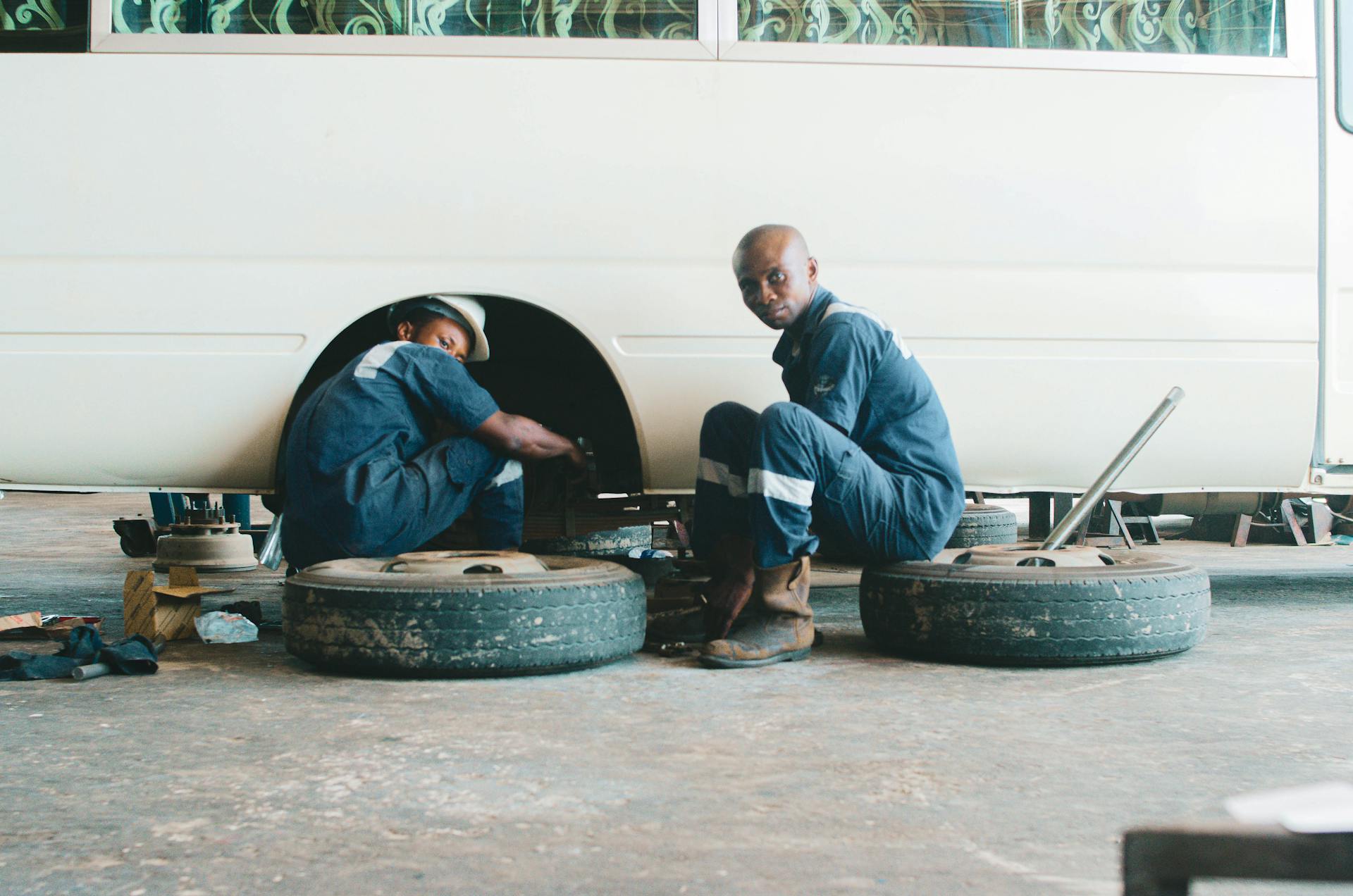 Two mechanics in coveralls repairing vehicle in Nigerian workshop, focusing on wheel maintenance.