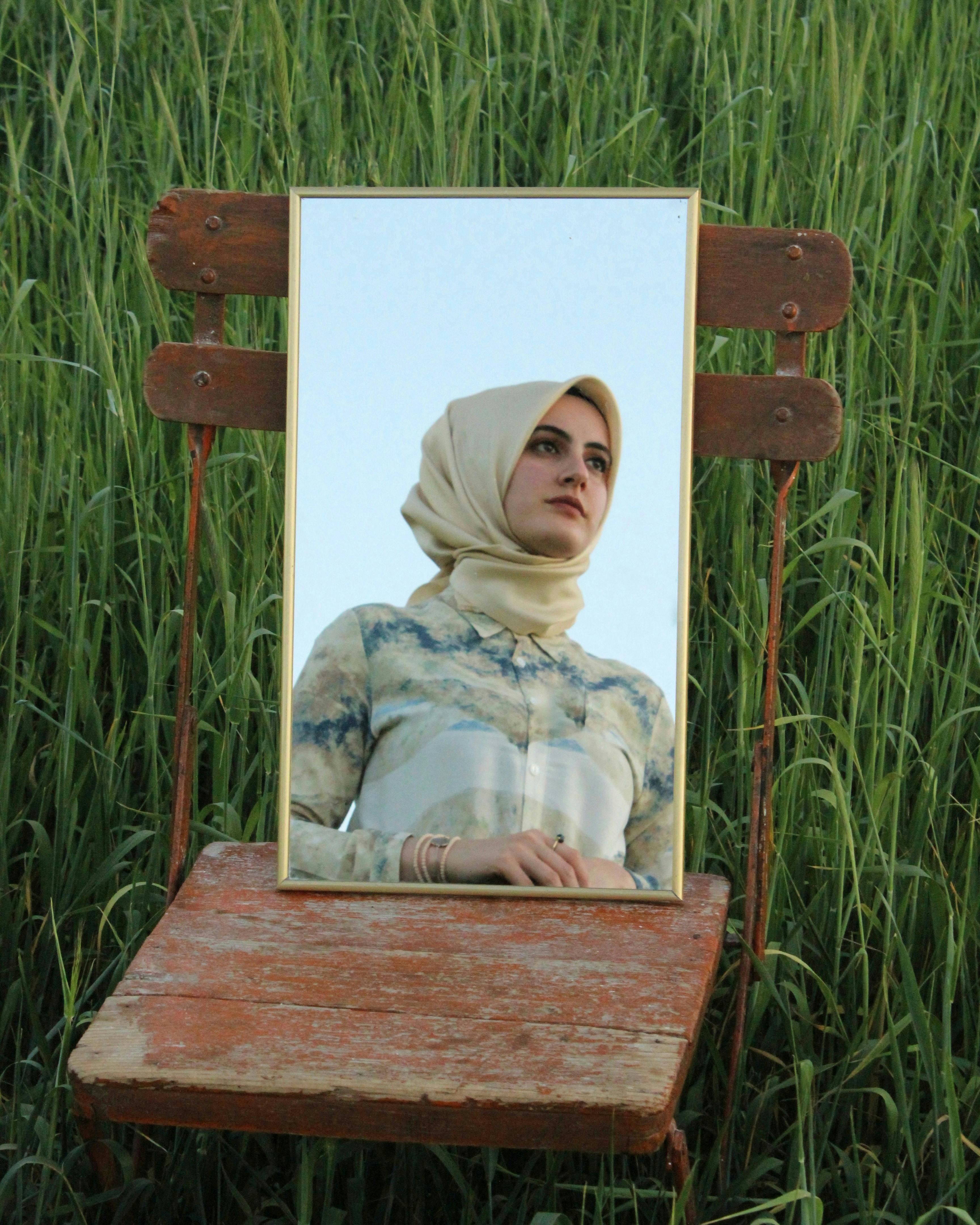 A woman in a headscarf is reflected in a mirror on a wooden chair in a grassy field.