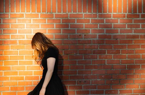 Free A woman in a black dress stands by a sunlit brick wall, casting a shadow. Stock Photo