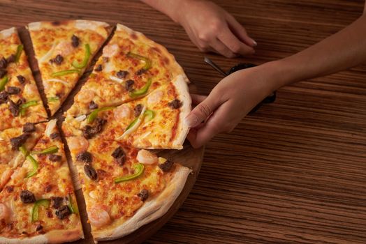 Hands reaching for a delicious pizza topped with shrimp, peppers, and beef on a wooden table.
