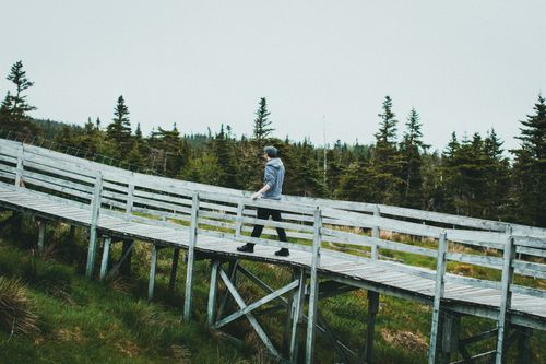 Free A solitary man walks across a rustic wooden bridge surrounded by serene forest scenery. Stock Photo