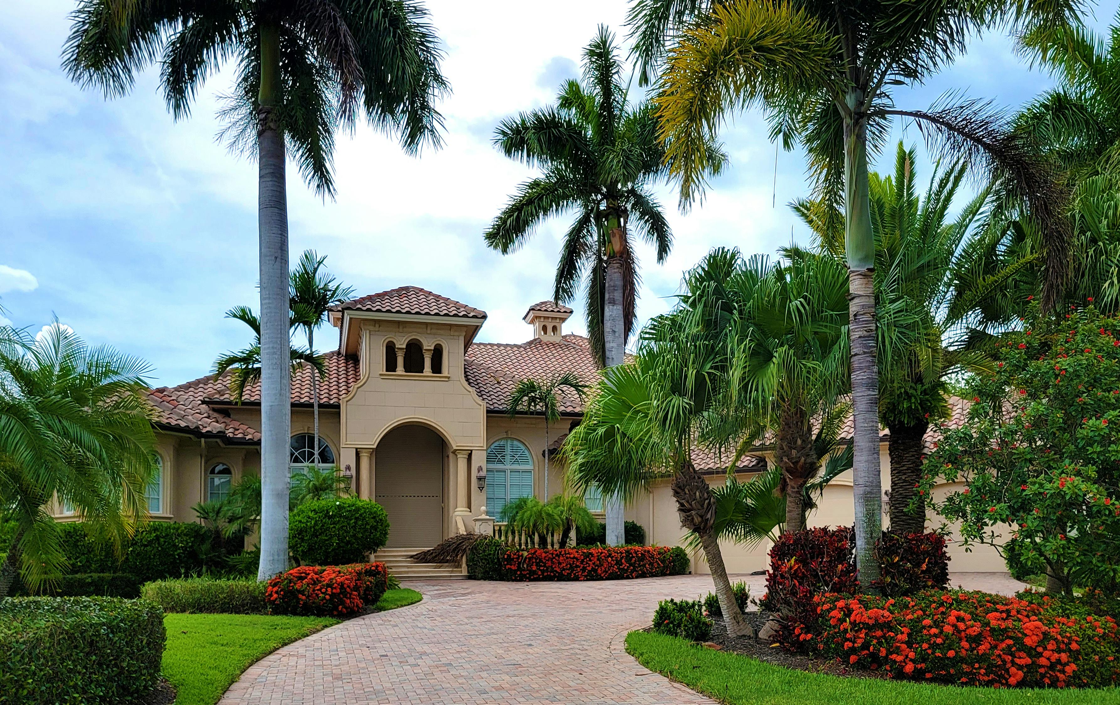 A large, beige house with a curved driveway, palm trees, and colorful flowers in a tropical setting.