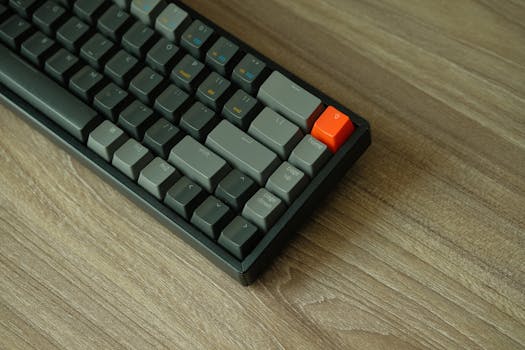 A detailed view of a mechanical keyboard with orange accent key on a wooden table.
