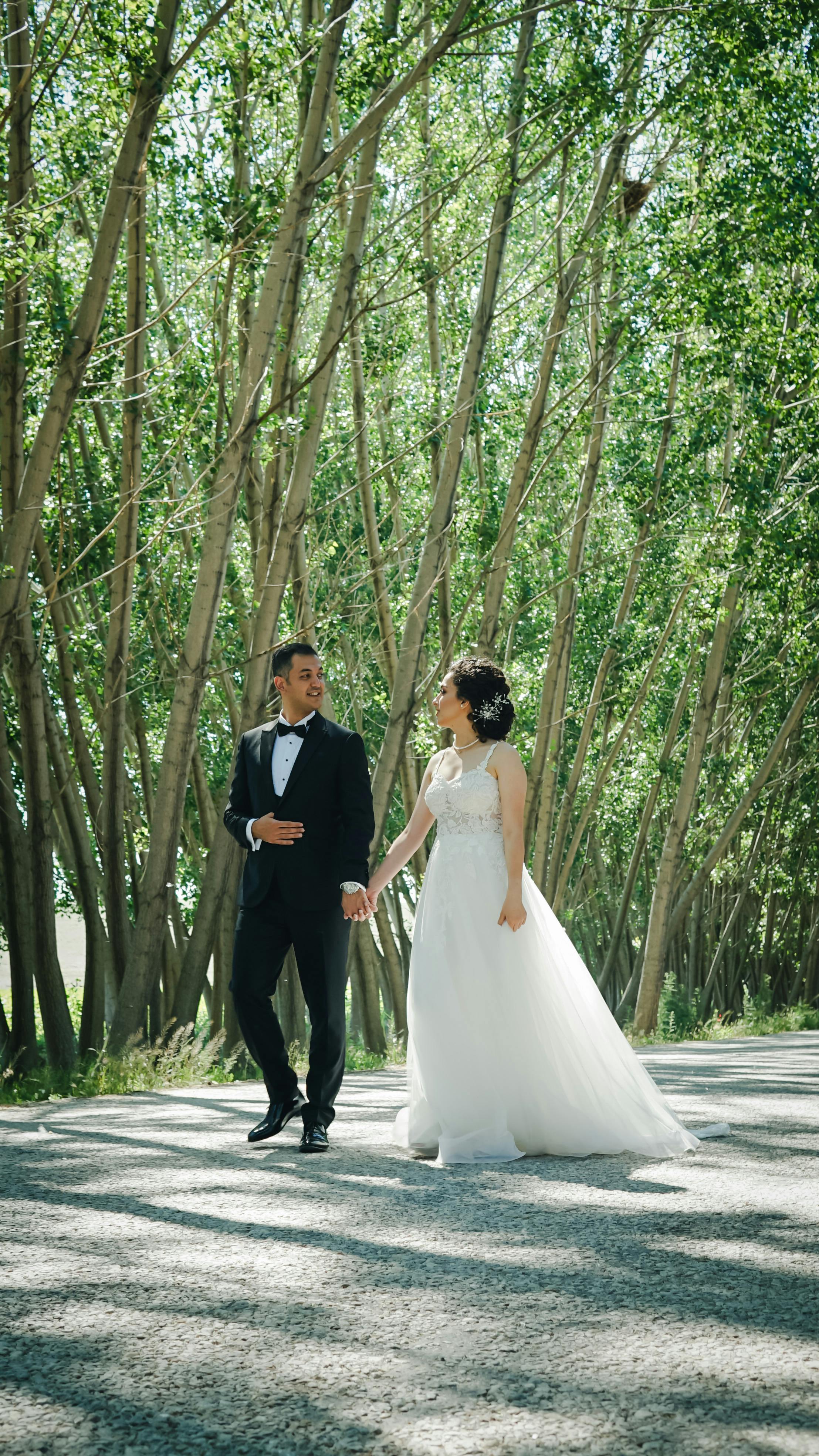 Bride and groom walking hand in hand under lush trees on their wedding day, showcasing love and elegance.