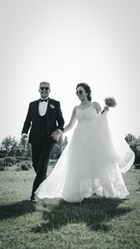 Bride and groom joyfully holding hands in a sunlit grass field, captured in black and white.