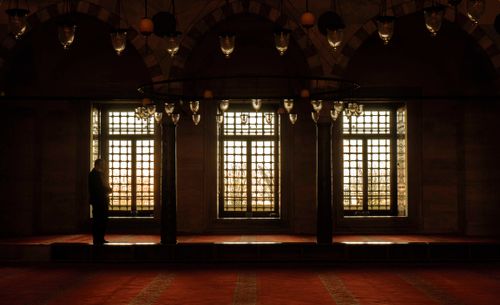Free Silhouette of a person standing in a dimly lit mosque with large decorative windows. Stock Photo