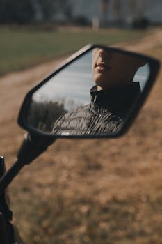 Artistic capture of a person's reflection in a motorcycle mirror along a rural road.