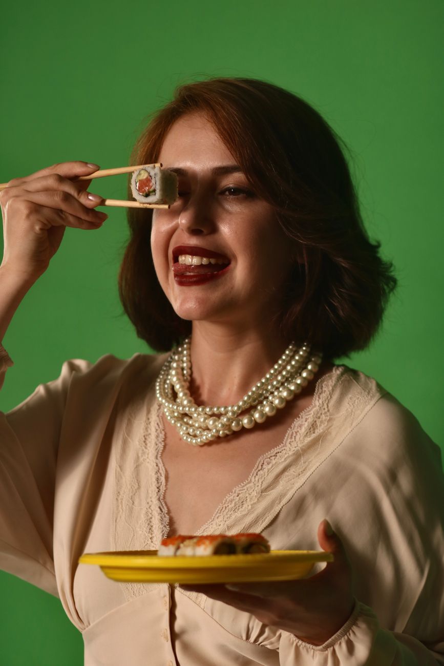 Smiling woman holding sushi with chopsticks, wearing pearl necklace.