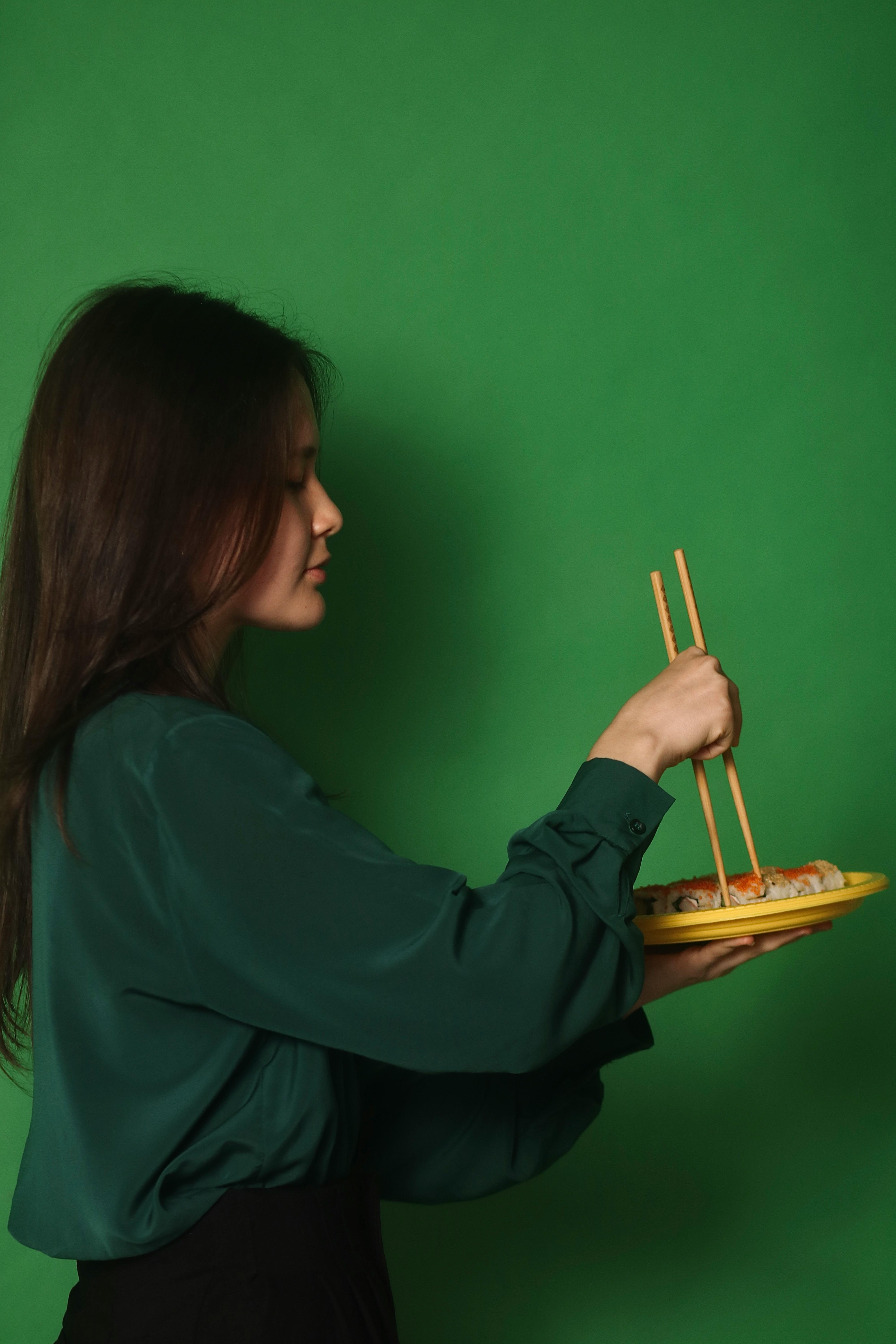 A woman holding a plate of sushi with chopsticks against a green background.
