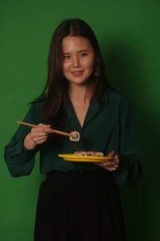 Asian woman holding sushi with chopsticks on a green backdrop.